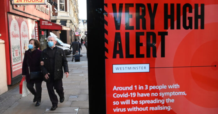 Ζευγάρι σε δρόμο του Λονδίνου δίπλα σε πινακίδα που προειδοποιεί για την έξαρση του Covid-19