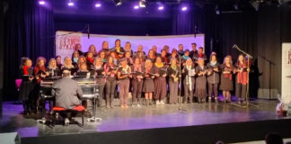 Πολυμελής χορωδία του Δήμου Θήρας στη σκηνή του ΕΠΑΛ Πάρου και ο μαέστρος κάθεται στο πιάνο